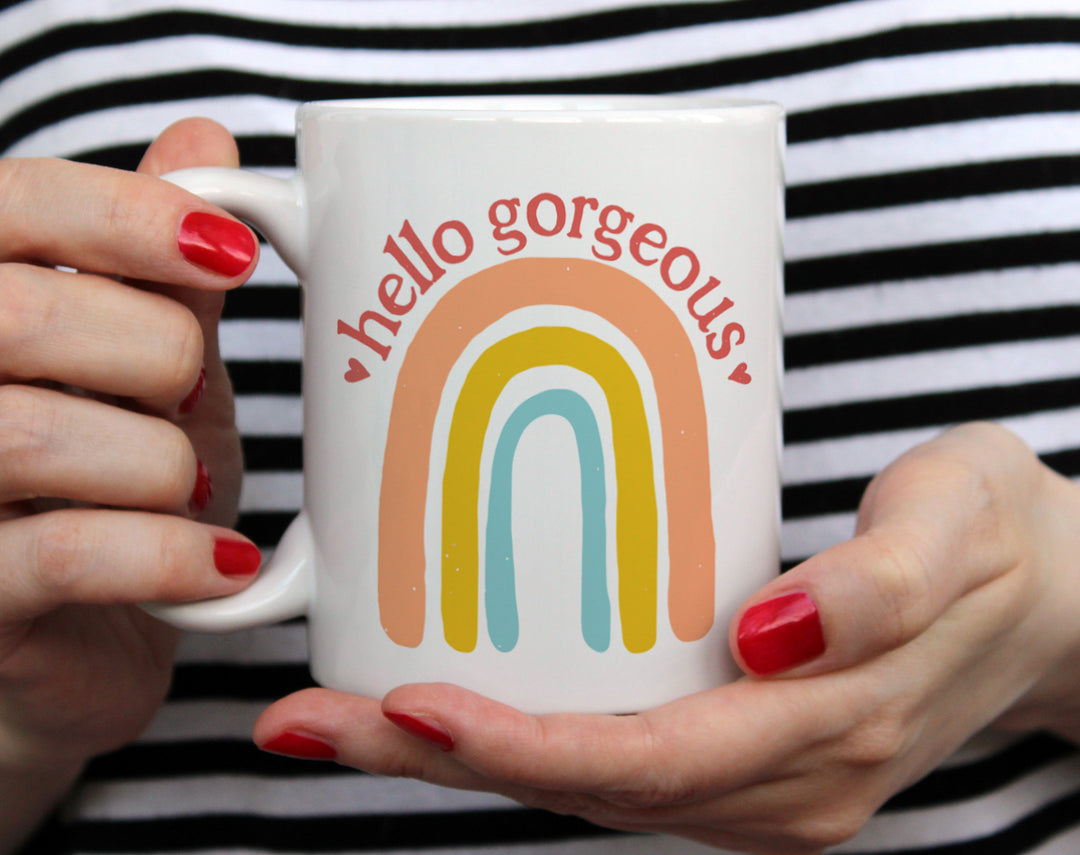 white mug that says Hello Gorgeous with a rainbow being held by woman wearing black and white top with red nail polish