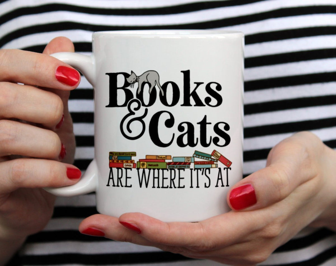 Books & Cats are Where It's At Mug held by a woman in black and white top with red nail polish
