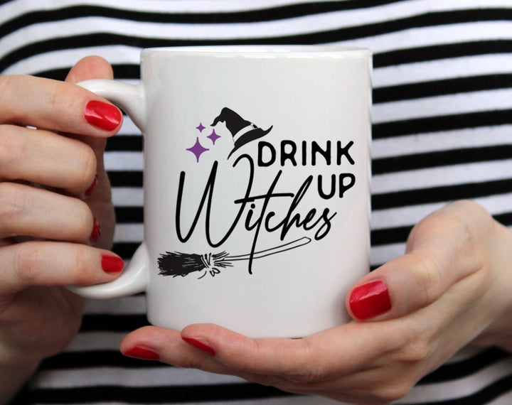 white mug that says Drink Up Witches with broomstick, witches hat, and three purple stars being held by woman wearing black and white top with red nail polish