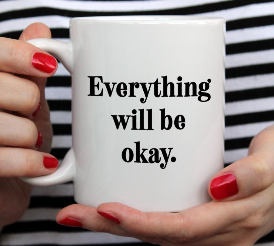 White mug that says Everything will be okay being held by woman wearing black and white top with red nail polish