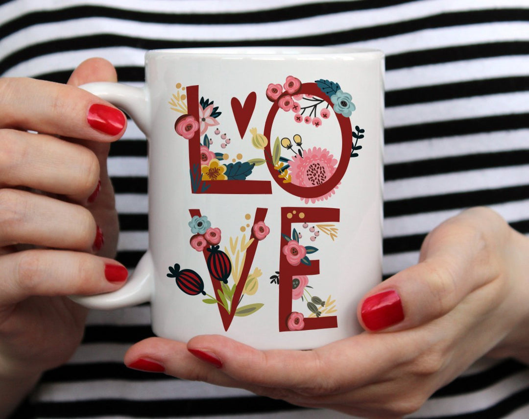 Mug that says Love with flowers around the letters being held by woman wearing black and white top with red nail polish