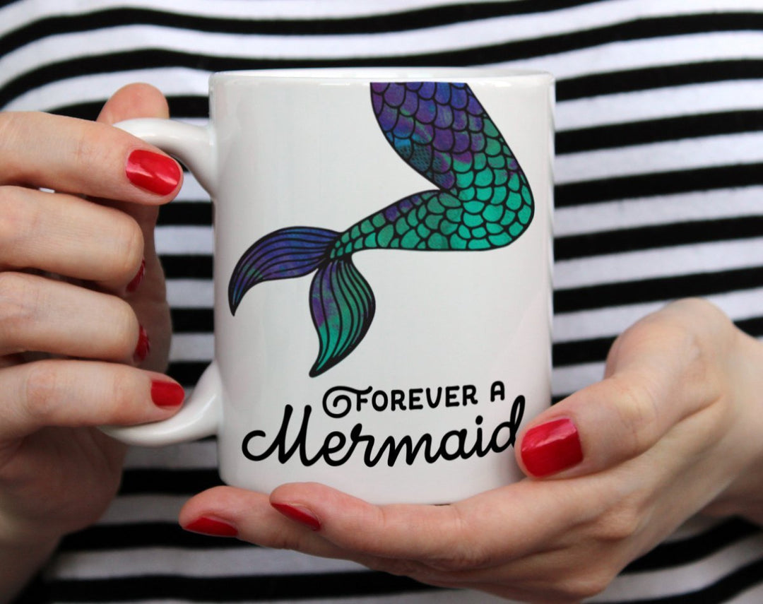 white mug that says Forever A Mermaid with a colorful mermaid tail on it being held by woman wearing black and white top with red nail polish