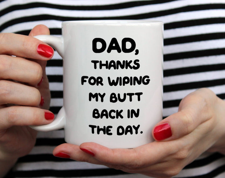 White mug that says Dad, thanks for wiping my butt back in the day being held by woman wearing black and white top with red nail polish