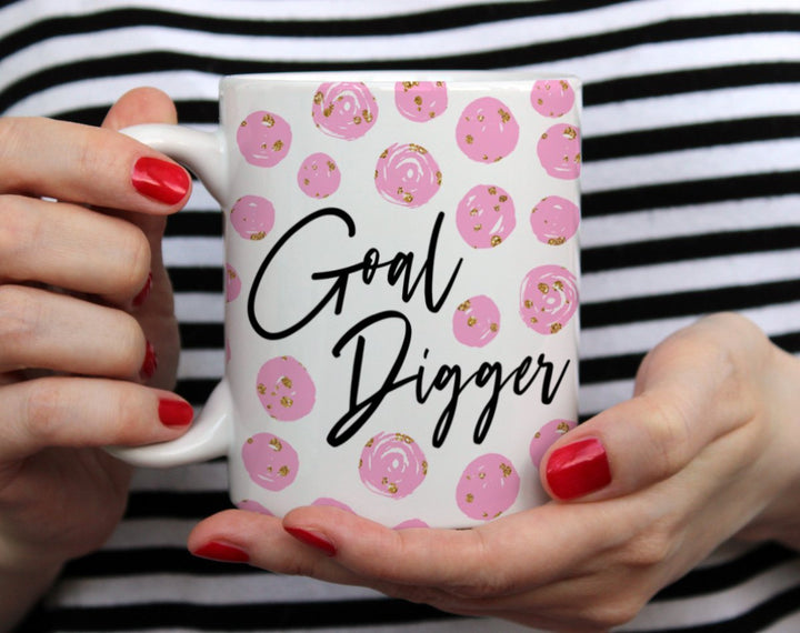 White mug that says Goal Digger with pink and gold spots on a white background being held by woman wearing black and white top with red nail polish