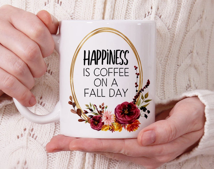 White mug that says Happiness is Coffee on a Fall Day with flowers being held by person