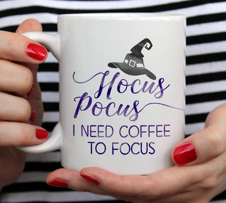 White mug that says hocus pocus I need coffee to focus with black witches hat being held by woman wearing black and white top with red nail polish