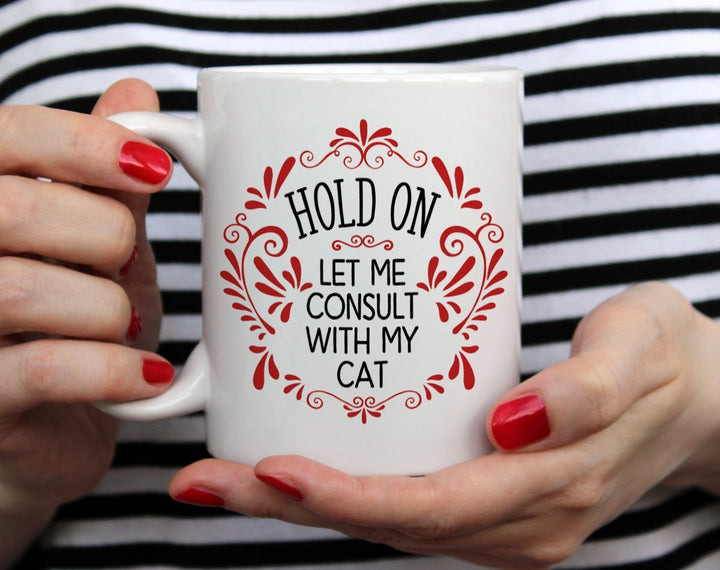 Mug that says Hold On Let me Consult with my Cat with red doodles being held by woman wearing black and white top with red nail polish