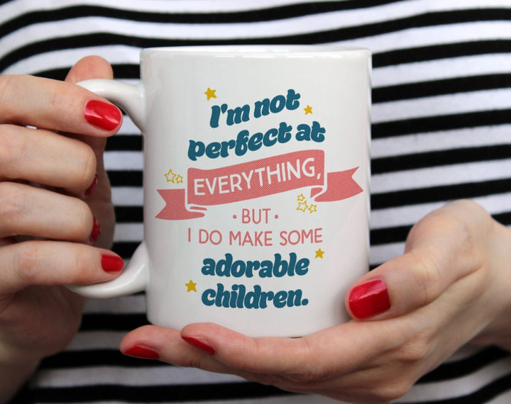 white mug that says I make adorable children mug with stars on it being held by woman wearing black and white top with red nail polish