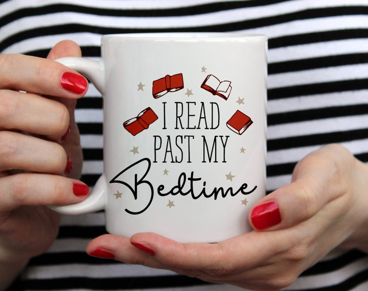 Mug that says I read past my bedtime with books floating around it being held by woman wearing black and white top with red nail polish