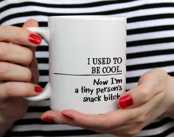 Mug that says I used to be cool, now I'm a tiny person's snack bitch being held by woman wearing black and white top with red nail polish
