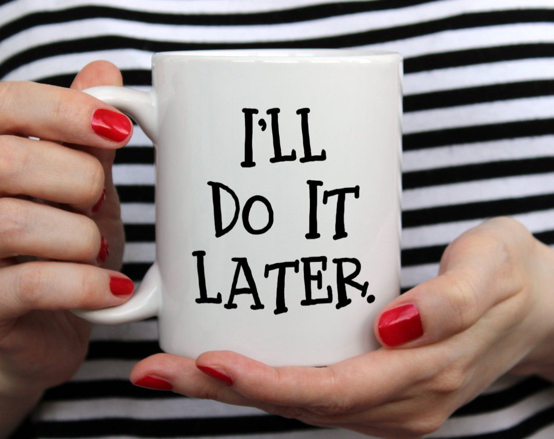 White mug that says I'll do it later being held by woman wearing black and white top with red nail polish