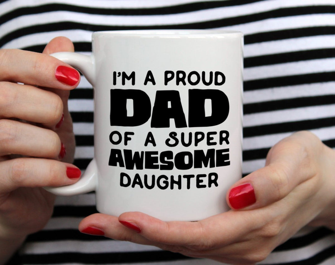 Mug that says I'm a proud dad of a super awesome daughter being held by woman wearing black and white top with red nail polish