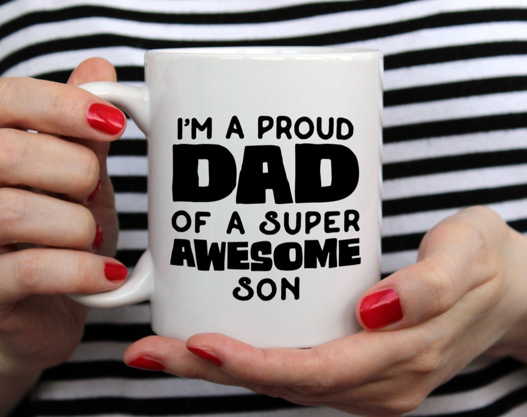 White mug that says I'm a proud dad of a super awesome son being held by woman wearing black and white top with red nail polish