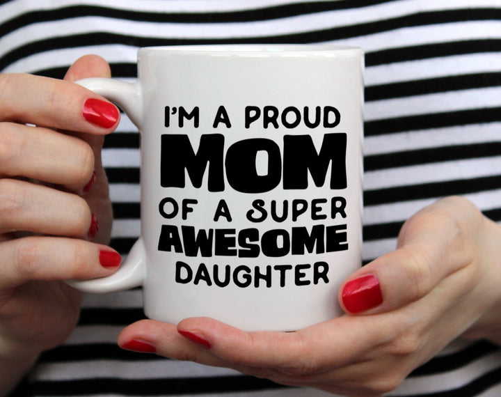 White mug that says I'm a proud mom of a super awesome daughter being held by woman wearing black and white top with red nail polish