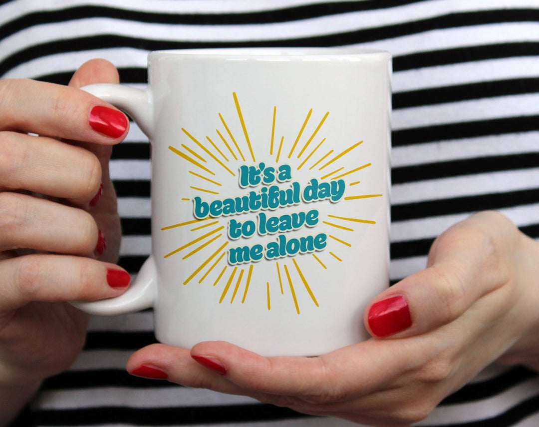 Mug that says It's a beautiful day to leave my alone with yellow bursts being held by woman wearing black and white top with red nail polish