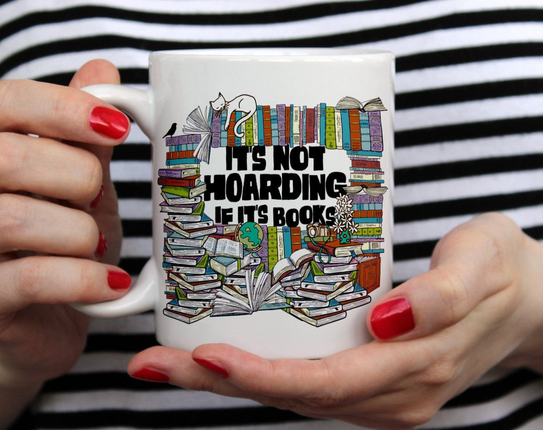 white mug that says It's Not Hoarding if it's Books with lots of books surrounding the words being held by woman wearing black and white top with red nail polish
