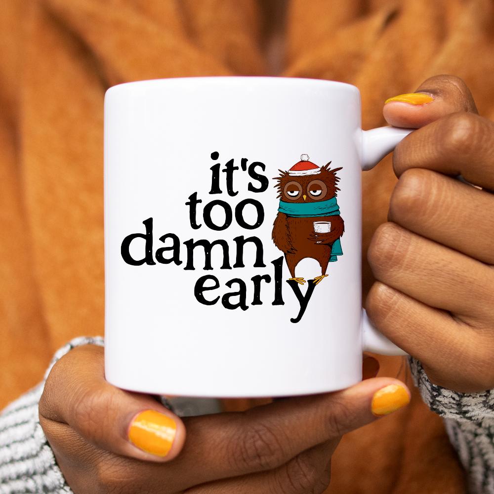 white mug that says It's Too Damn Early with an owl drinking coffee being held by person with yellow nail polish