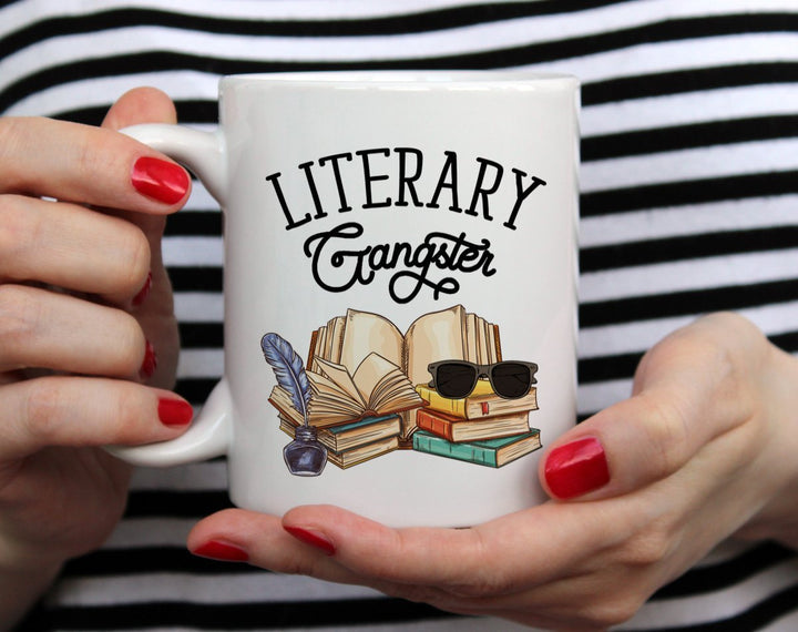 White mug that says Literary Gangster with books and sunglasses being held by woman wearing black and white top with red nail polish