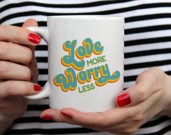 White mug that says Love More, Worry Less in blue and yellow text being held by woman wearing black and white top with red nail polish